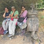Three walkers share a bench with a carved wooden sculpture of a monk, resting with eyes closed.