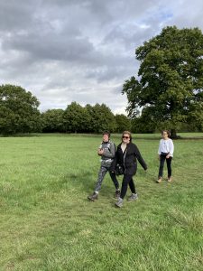 Three walkers cross a green field, there are mature trees in green leaf in the background.