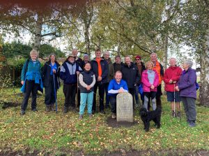 Fifteen walkers on a village green. The trees above are losing their leaves creating a carpet of autumn colours. The group are gathered around a stone waymarker for the North Downs Way national trail.