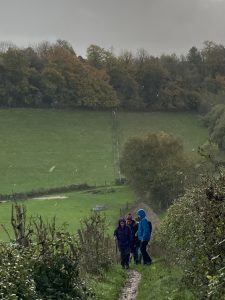A group of walkers in rain jackets ascend a steep path between hedgerows. There are green fields in the valley and woodland behind. Skies are grey.