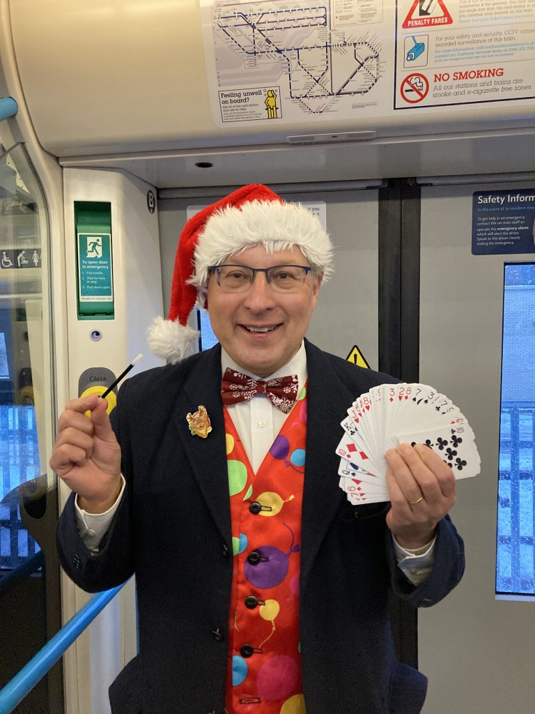Magi Paul in his festive waistcoat and santa hat. He is holding a fanned-out deck of cards and a magic wand