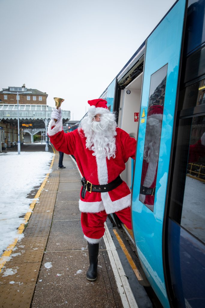 Santa at the doors to a blue train at the station. Santa has one foot on board, the other on the platform and is ringing a golden bell to signal the train is ready to depart. The station latform is covered with snow.