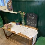 A small wooden table with a white table cloth. On the table are a welcoming plate of biscuits, a vase of flowers, a box of pens and survey forms to complete. A chalk board sign invites people to "Share your views, fill in a survey!!"