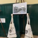 A wooden easel displaying a black and white photo of a steam engine at Hollingbourne station. Two cotton tote bags advertising Kent Community Rail Partnership are hanging from the easel with a "Hop on board the train" logo which includes an image of hops