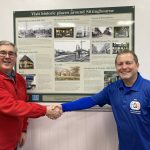 Richard Emmett (wearing red) and Gary Outram (wearing blue) smiling and shaking hands in fromt of the local history board in the waiting room.