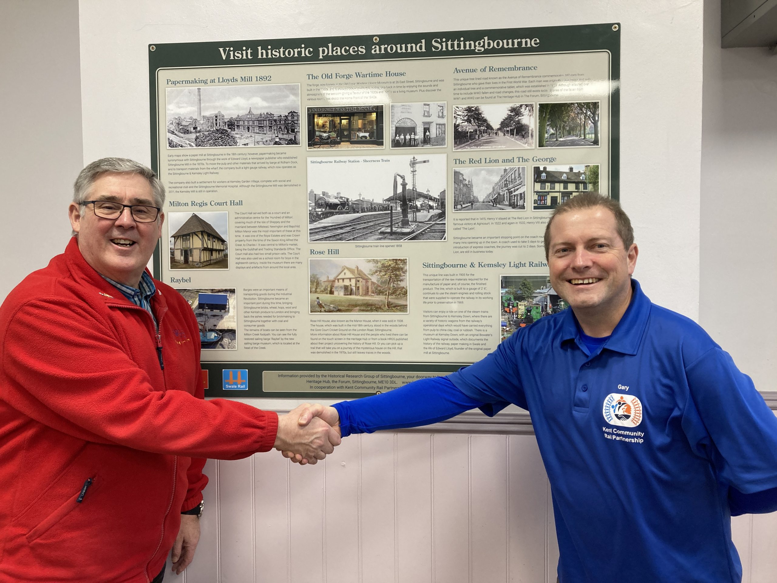 Richard Emmett (wearing red) and Gary Outram (wearing blue) smiling and shaking hands in fromt of the local history board in the waiting room.