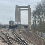 A Southeastern train approaching Swale station over the imposing concrete structure of the Kingsferry lifting bridge. The sky is grey.
