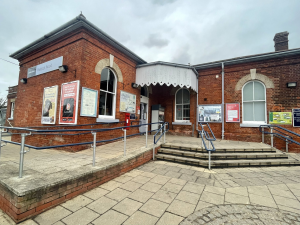The entrance to Paddock Wood station, a paved forecourt with steps and a ramp leads to the doors of a red-brick victorian station building. A small white wooden canopy provides shelter to the entrance door.