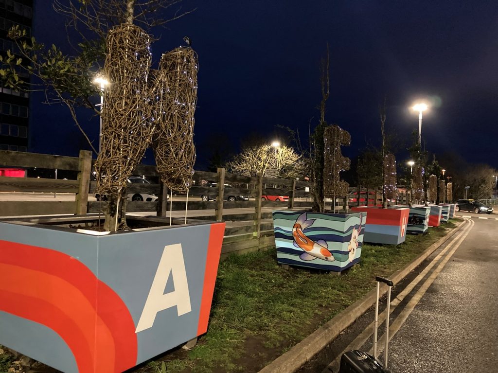 A night time scene. Colourful planters featuring flowing shapes and fish spell out "Ashford". Mounted in the planters are willow sculptures that spell "Welcome"