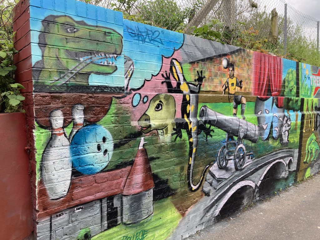 Colourful mural panel featuring a dinosaur, bowling alley, hop farm, lizard, cannon, bridge, theatre curtains.