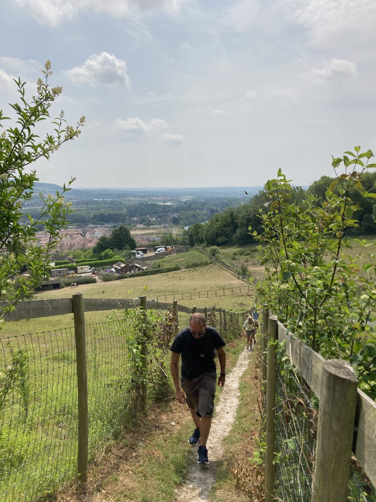 Walkers climb a steep unmade path between fields. The view stretches into the distance past houses to green hillsides, the sky is bright but cloudy.