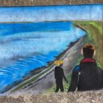 A painted mural of two people walking along the seafront. The sky and sea are blue, a pebble beach with a green pathway ahead