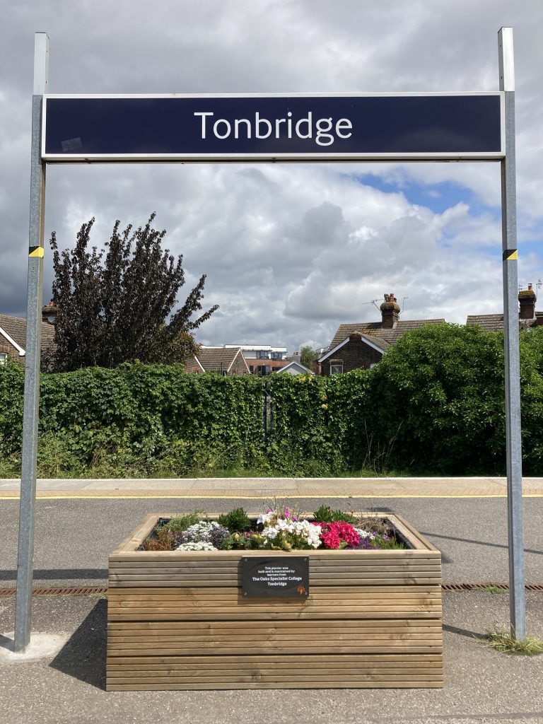 A wooden planter filled with coloufrul flowers beneath a sign for Tonbridge station.