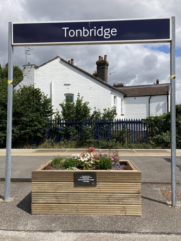 A wooden planter filled with coloufrul flowers beneath a sign for Tonbridge station.