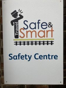 Sign for Travel Safe & Travel Smart Safety Centre. Logo features an image representing a member of platform staff calling out a yellow platform edge marking and a rail track.