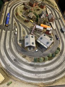 A model railway layout with platforms, sidings, level crossings, a blue engine, model cars, peoples and buildings. The model depicts a variety of poor and unsafe behaviours around the railway and is used in a "how many dangers can you see" context.