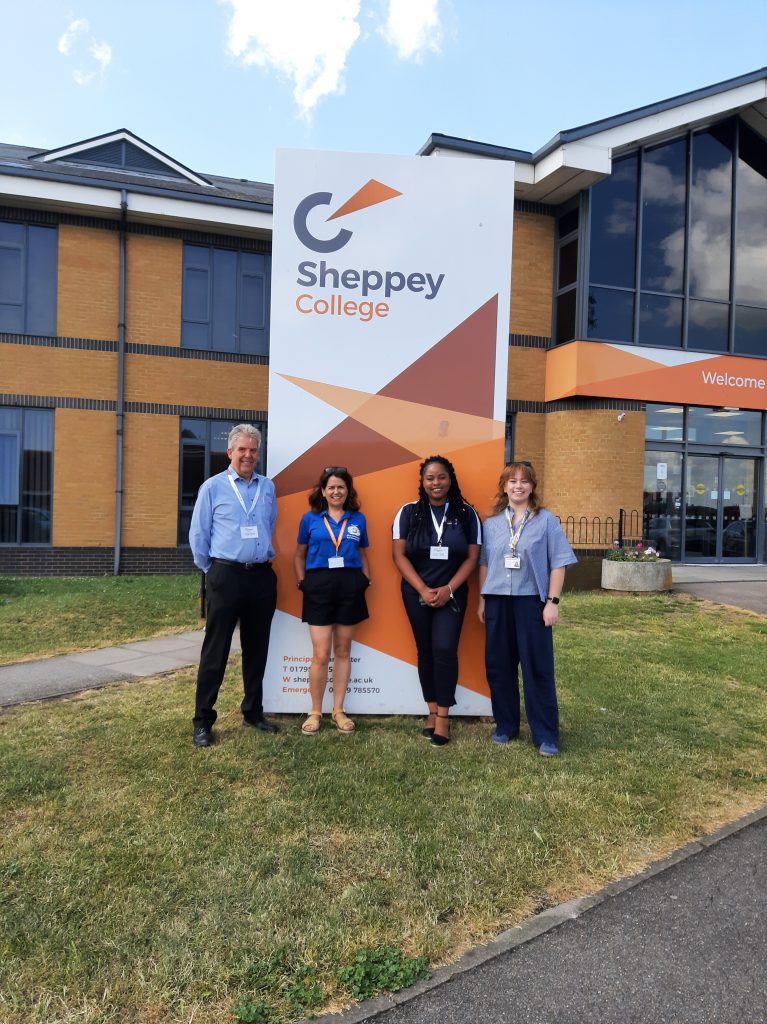 Four people beneath a large totem sign for Sheppey College