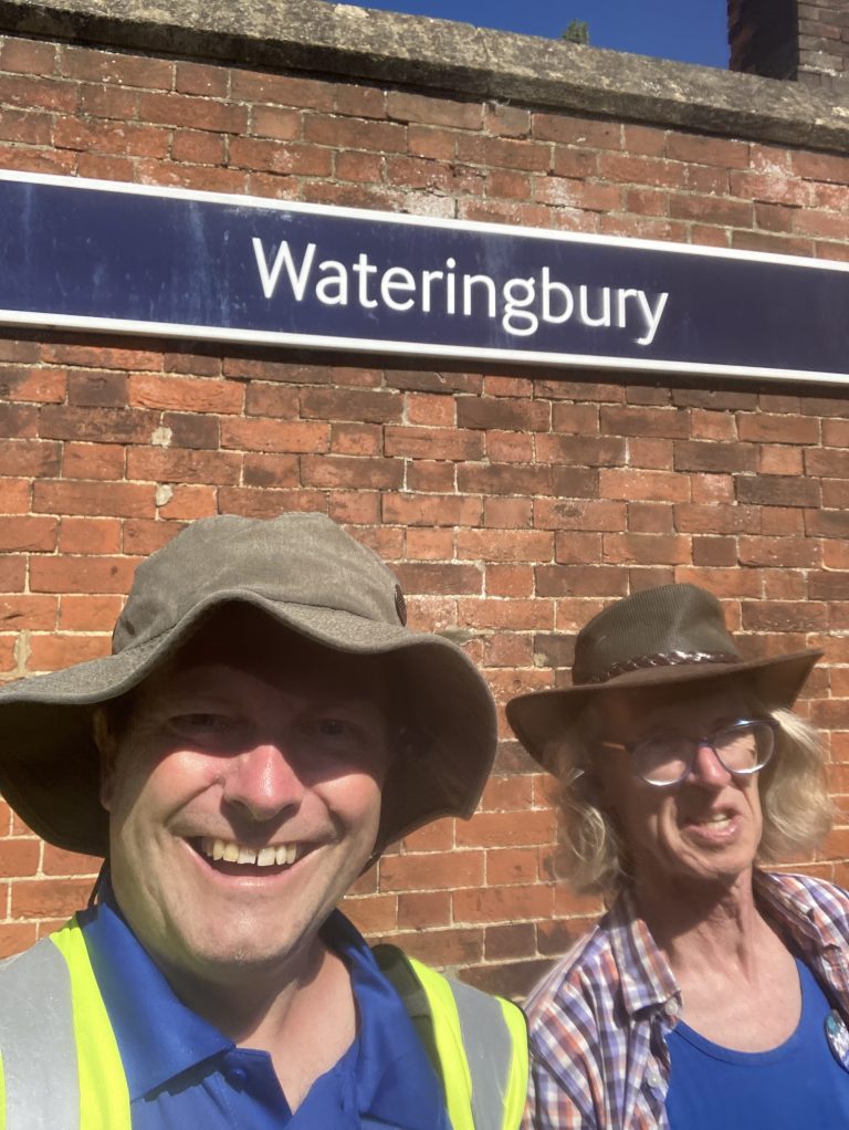 Dave and Gary in wide-brimmed hats in front of the Wateringbury station sign