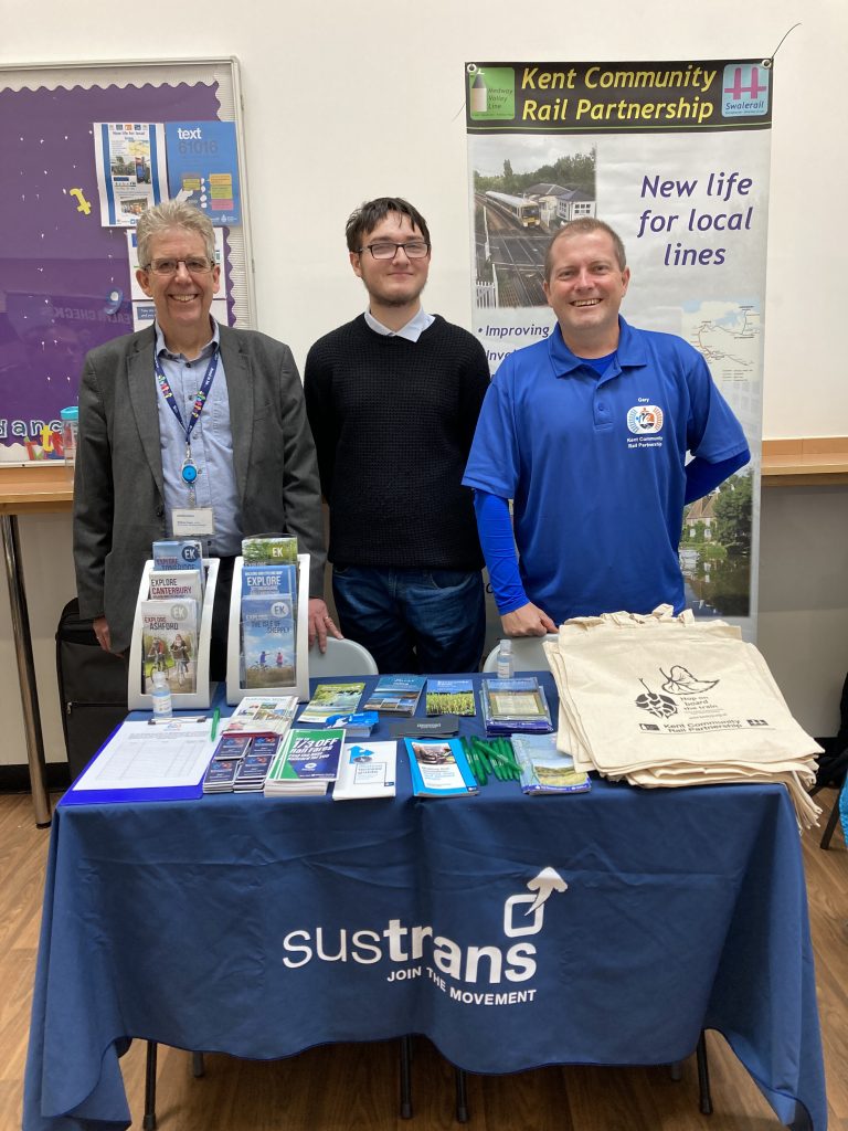 Matt, Dominic and Gary standing at our display stall. In front of them is a table with tot-bags, pens, fridge magnets and a variety of brochures promoting railcards, assisted accessibility services and local walks. Behind them are banners and posters promoting Kent CRP, British Transport Police 61016 and "Go Green By Train" messaging
