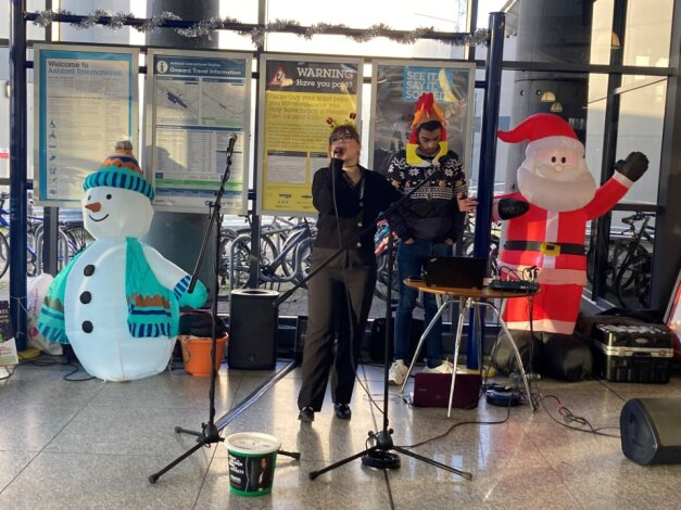 Two people singing in a station foyer, behind them are an inflatable snowman and Santa.