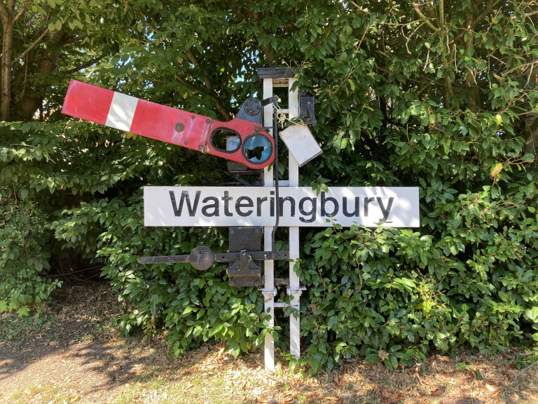 A semaphore railway signal and station name board "Wateringbury" mounted on a set of short poles just a few feet above the ground. Behind is a thick green hedge.