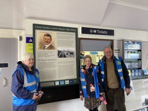 Three people pictured in the booking hall at Queenborough station. Behind them are the ticket office window and a new information panel outlining the story of Colonel Stephens and the Sheppey Light Railway