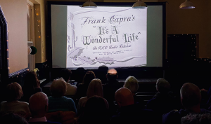 A crowd seated infront of a projector screen, the opening title of "It's a Wonderful Life" is displayed.