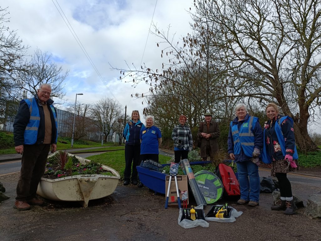 A group of people standing by two rowing boats that are used as flowerbeds. In front are a variety of pieces of new gardening equipment being presented to the group.