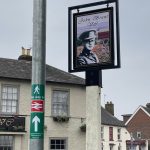A green waymarking sticker with British Rail double arrow symbols attached to a lamppost. Adjacent is the pub sign of the John Brunt VC, displaying an image of Captain John Brunt.