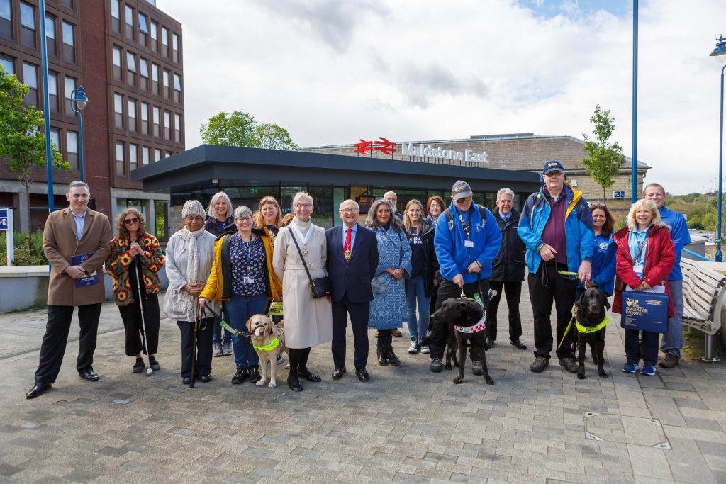 A large group of people and guide dogs posing for a group photo in front of the entrance to Maidstone East station.