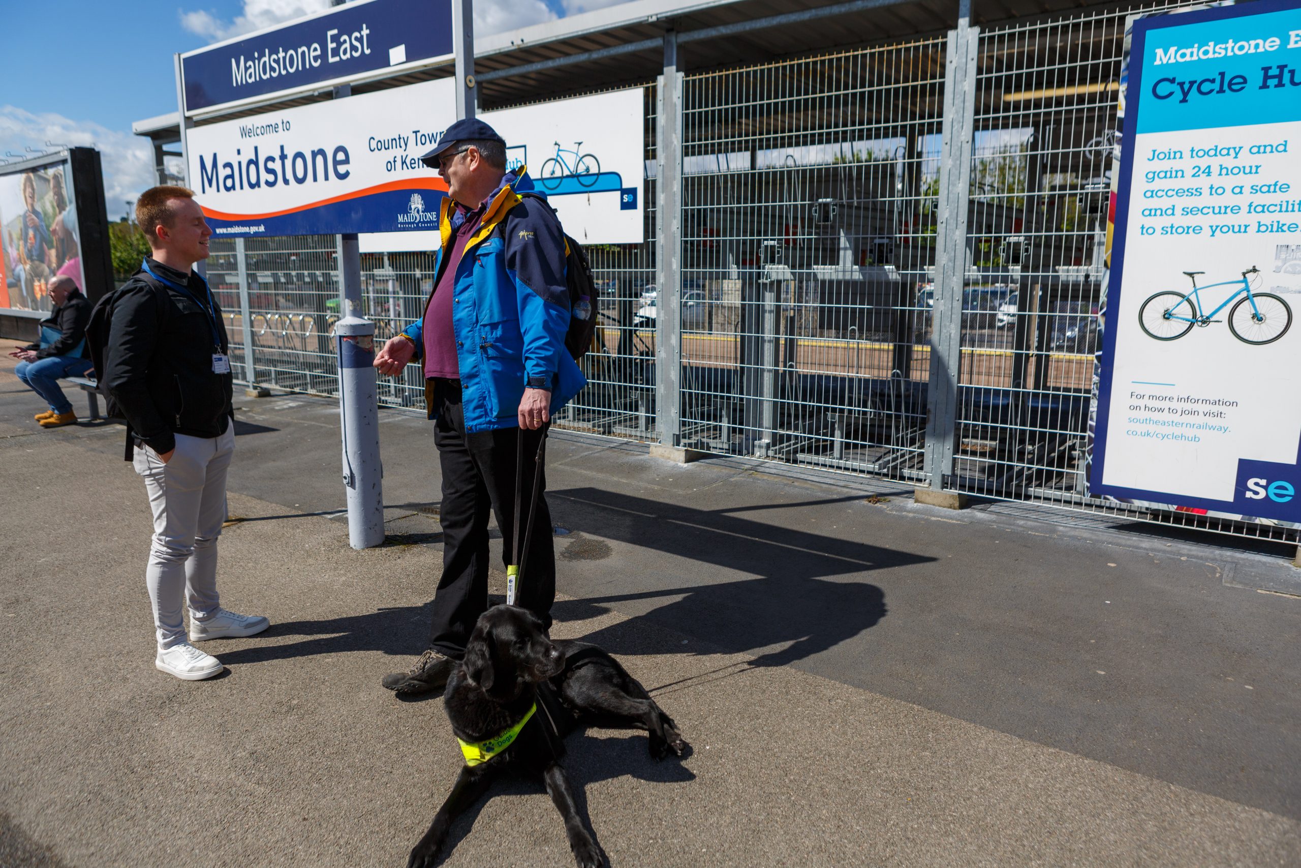 A guide dog owner and their guide dog chatting on the platform. Behind them is a secure cycle hub.