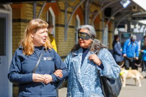 A sighted guide leads a blindfolded volunteer along a station platform. In the background are more event participants and their guide dogs