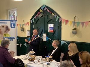 The Mayor of Maidstone wearing his chain of office is stood by a Guide Dogs banner poster, speaking to the audience. Afternoon tea is set out on a table in front of the mayor and other guests