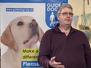 A guide dog user stood by a Guide Dogs banner display presents to the audience