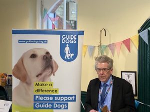 Matt Fraser stood beside a Guide Dogs banner display, presenting to the room from notes on his phone
