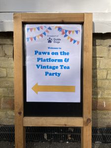 An A-Frame sign bearing a poster "Guide Dogs, Welcome to Paws on the Platform and Vintage Tea Party"