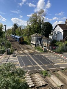 View from a footbridge of a blue liveried Southeastern train as it approaches a level crossing and white ship-lap boarded signal box