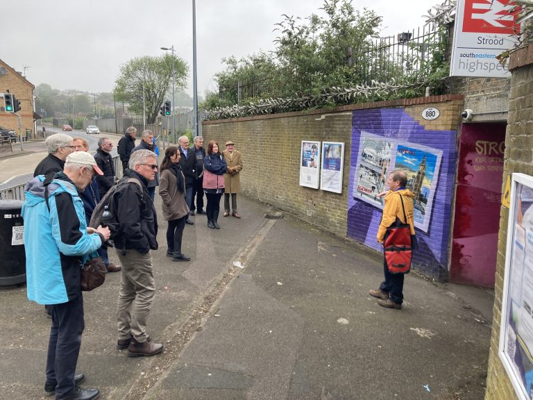 A crowd of people listening to Gary who is stood in front of some colourful murals at a subway entrance.