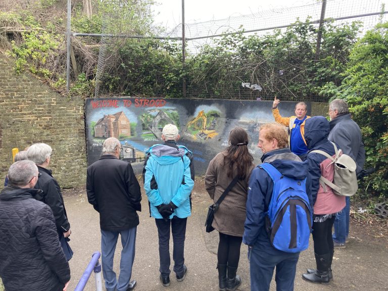 A crowd of people listening to Gary who is stood in front of some colourful murals at a subway entrance.