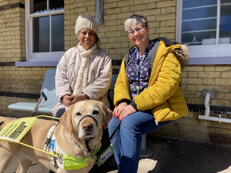 Two people seated on a bench on a railway platform. One is wearing a thick cream coloured coat, hat and scarf. There is a walking stick beside them. The other is wearing blue jeans, blouse and a padded yellow coat. In front of them is a golden coloured guide dog wearing an "on duty" reflective harness. Everyone is smiling.