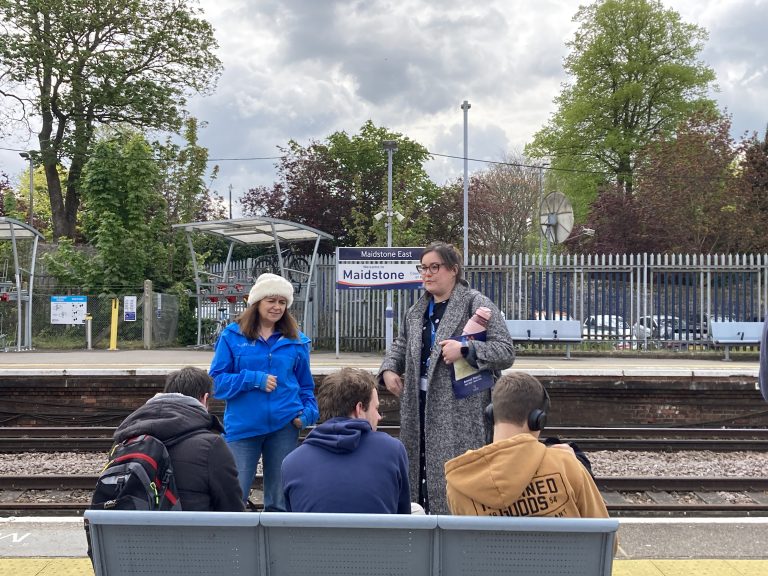 Students on a bench at the station listen to Therese and a tutor.