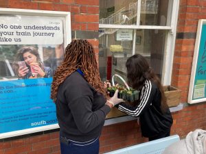 Two people with a caddy of small colourful flowers in pots begin to plant a windowbox. Either side of the window are posters, one promoting the Southeastern app, the other Samaritans helpline