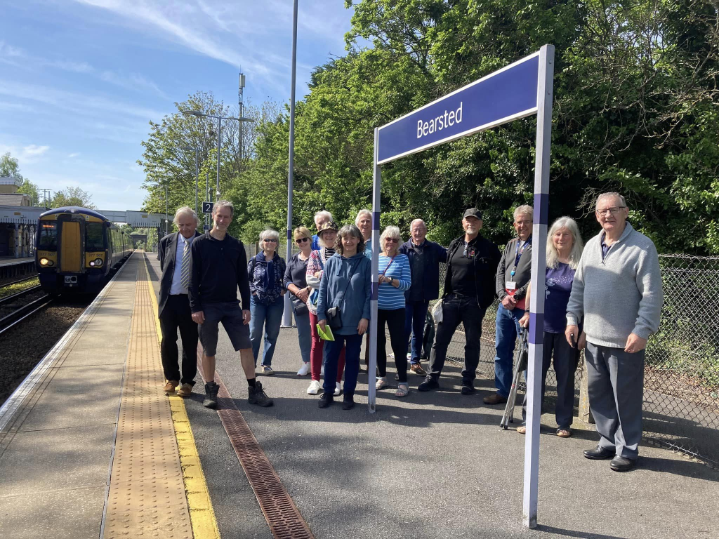 A crowd of people gathered on the platform at Bearsted station, near a station name sign. A train is arriving at the station