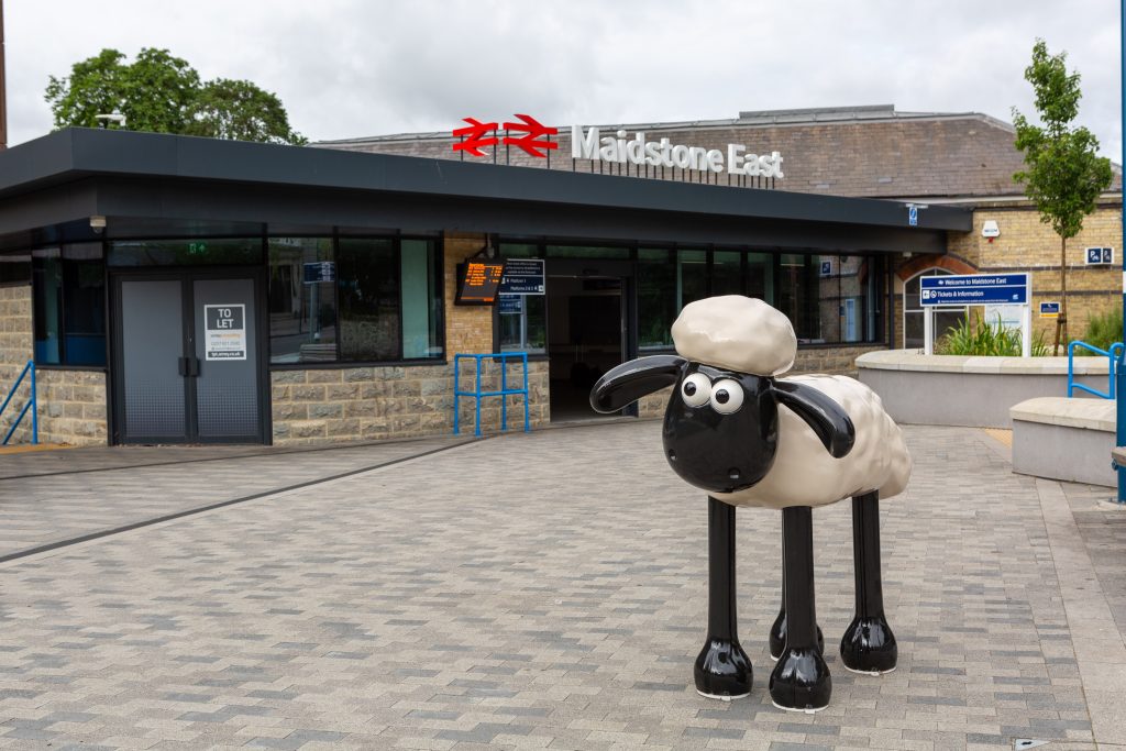 Shaun the Sheep outside Maidstone East station. A statute of a cartoon sheep, Shaun. Shaun has a black face, ears and legs and a fluffy white body and head. Shaun is stood on a paved forecourt area in front of a modern glass fronted station building. On the roof of the building are British Rail "double arrow" logos and the station name, "Maidstone East".