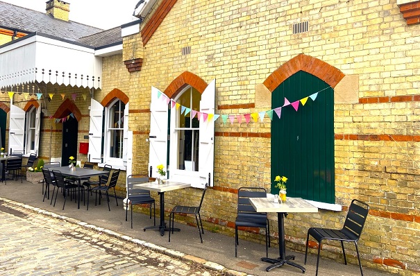 A brick built station building with shuttered windows. The outsides of the closed shutters are green, the insides of the open shutters are white. A white painted wooden canopy extendsabove the green entrance doors. There are tables and chairs outside with vases of yellow daffodils. Colourful bunting is strung between the windows and under the canopy.
