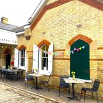 A brick built station building with shuttered windows. The outsides of the closed shutters are green, the insides of the open shutters are white. A white painted wooden canopy extendsabove the green entrance doors. There are tables and chairs outside with vases of yellow daffodils. Colourful bunting is strung between the windows and under the canopy.