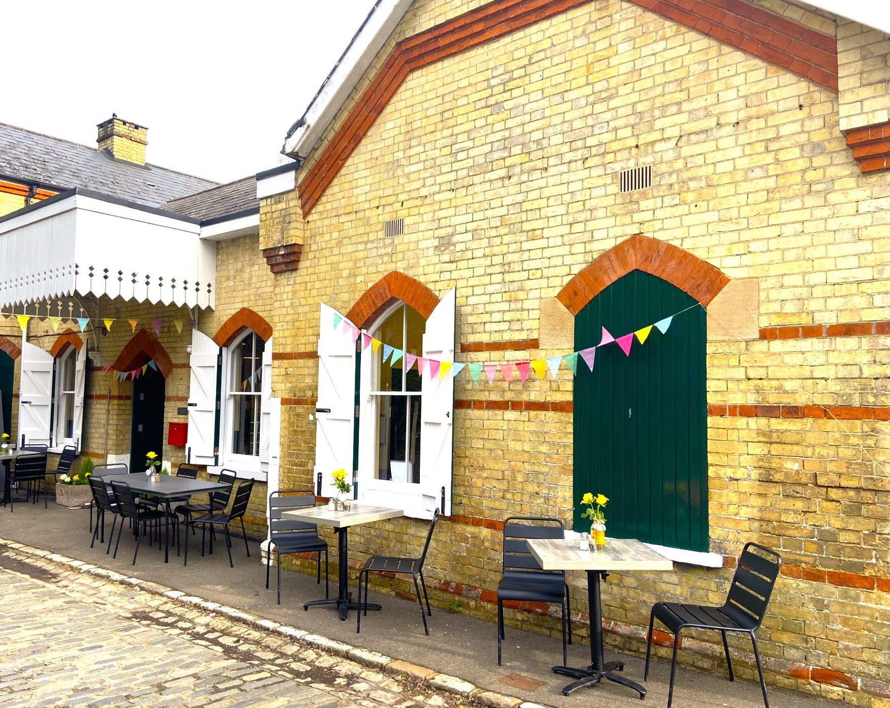 A brick built station building with shuttered windows. The outsides of the closed shutters are green, the insides of the open shutters are white. A white painted wooden canopy extendsabove the green entrance doors. There are tables and chairs outside with vases of yellow daffodils. Colourful bunting is strung between the windows and under the canopy.