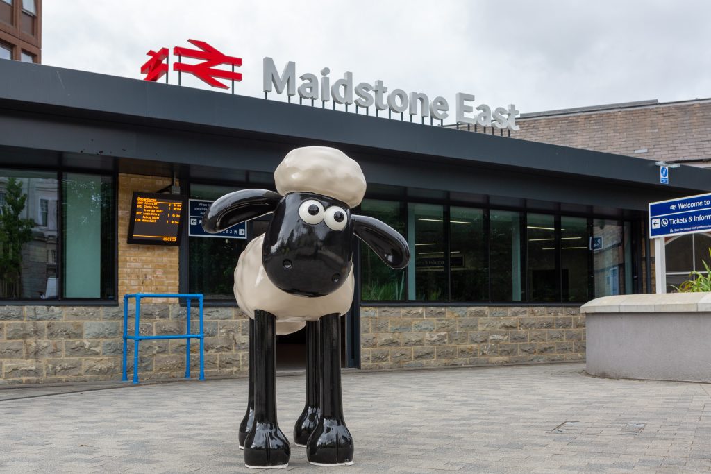 Shaun the Sheep outside Maidstone East station. A statute of a cartoon sheep, Shaun. Shaun has a black face, ears and legs and a fluffy white body and head. Shaun is stood on a paved forecourt area in front of a modern glass fronted station building. On the roof of the building are British Rail "double arrow" logos and the station name, "Maidstone East".