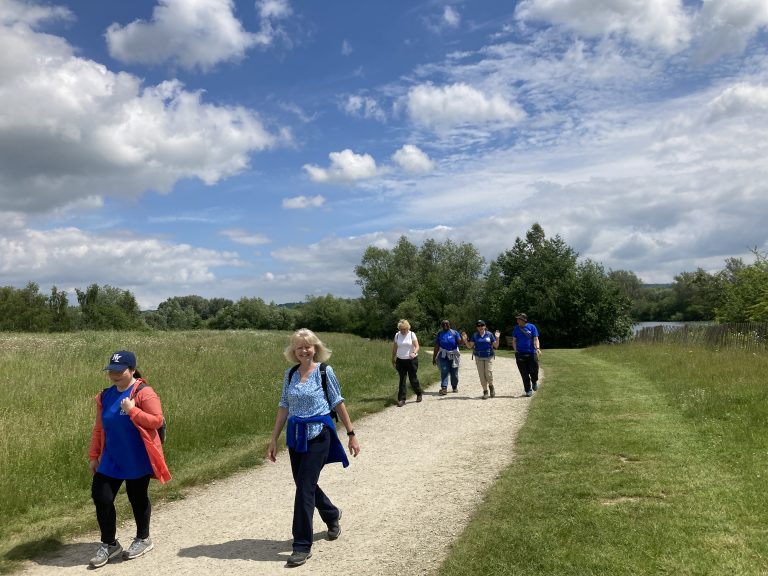 A group of walkers on a wide gravel path between freshly mown verges. There is a glimpse of a lake between trees and hedgerows in the background.