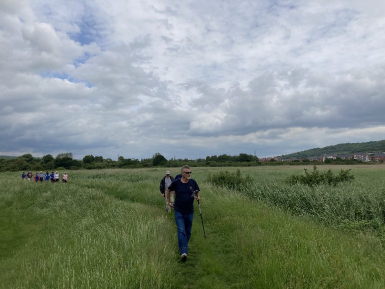 A group of walkers on a riverbank path lined with tal grass. A hedgerow follows the line of the river behind them, green hills rise in the distance. There are houses on the far bank of the river.